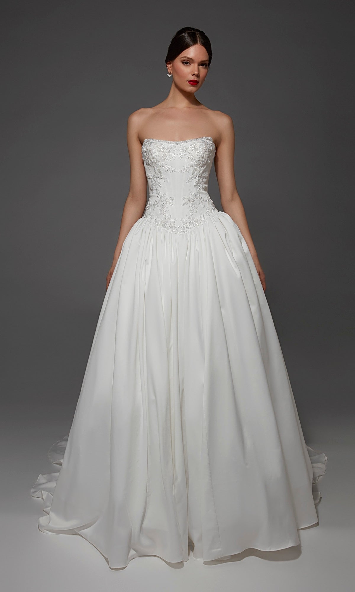 A woman wears the Alyce 7188 long wedding dress in Diamond White, featuring a strapless satin ball gown silhouette with floral embellishments on the bodice, standing against a dark gray background.